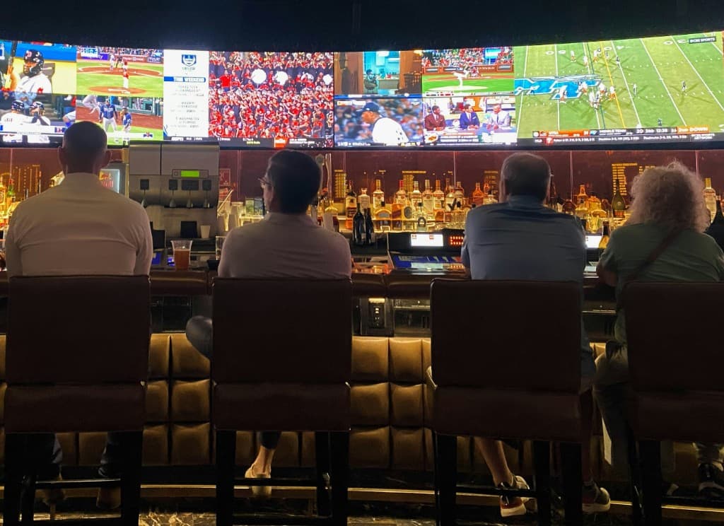Patrons seated at a sportsbook bar watching a wall of screens showing live games and posted lines.